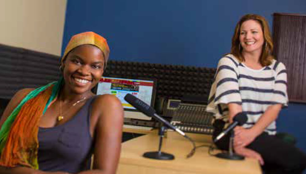 Two women sitting at a desk next to two microphones and a computer screen.