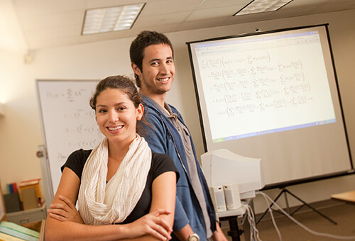 Photo of 2 students side by side inside a classroom with a whiteboard next to them.